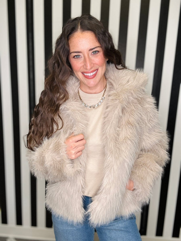 A woman with long wavy brown hair smiling, wearing a fluffy light beige faux fur jacket over a cream top and blue jeans, standing in front of a black and white vertically striped background.