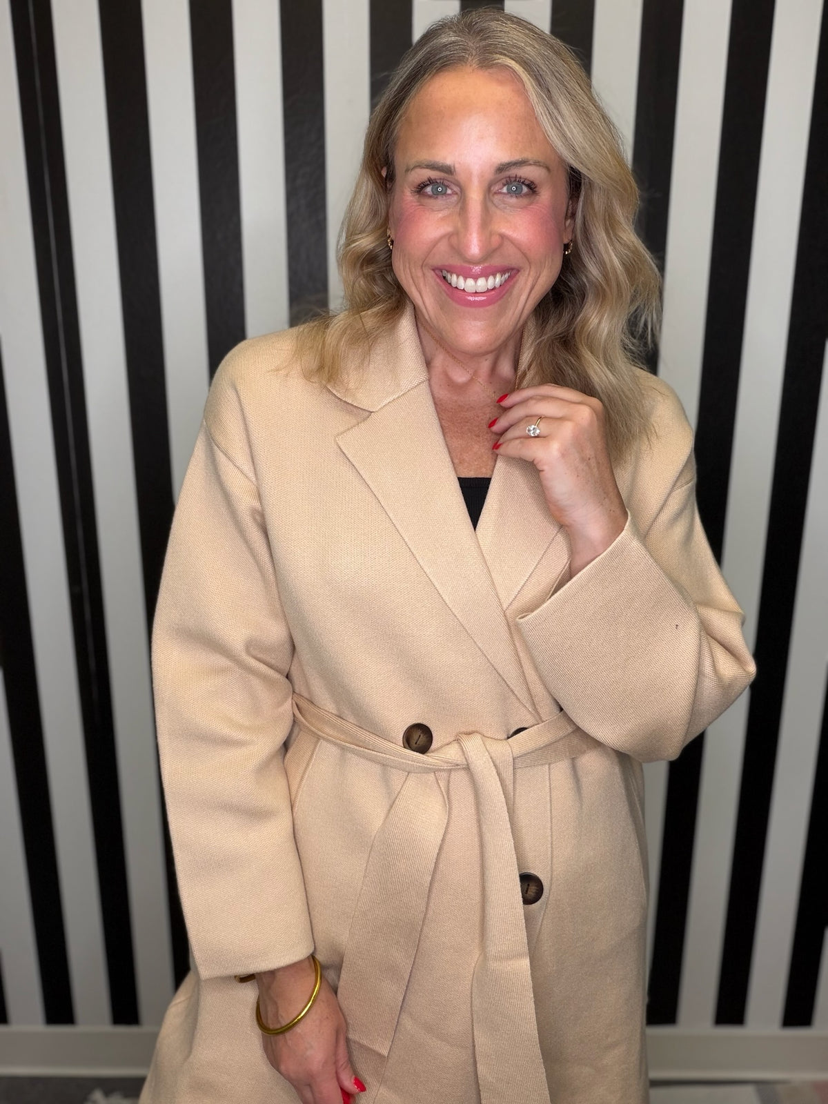 Woman in a brown trench coat standing in front of a black and white striped wall smiling.