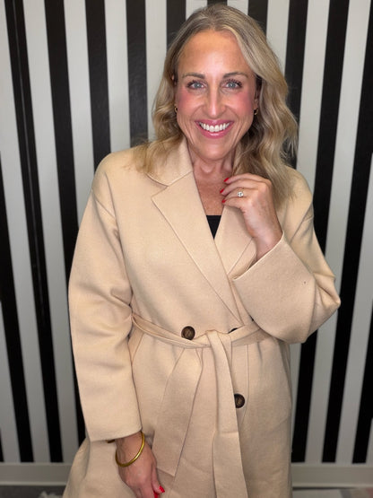 Woman in a brown trench coat standing in front of a black and white striped wall smiling.
