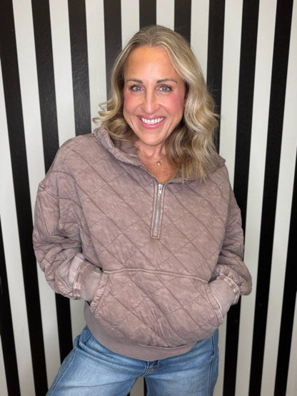 A smiling blonde woman wearing a taupe quilted half-zip pullover hoodie and blue jeans, standing in front of a black and white vertically striped wall.