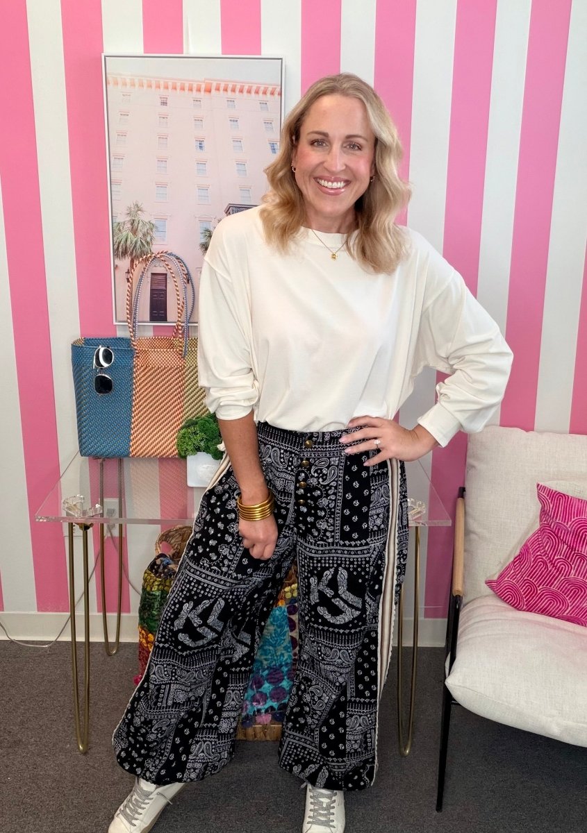 A woman posing in a white long-sleeve top and black-and-white patterned wide-leg pants, standing in front of a pink and white striped wall.