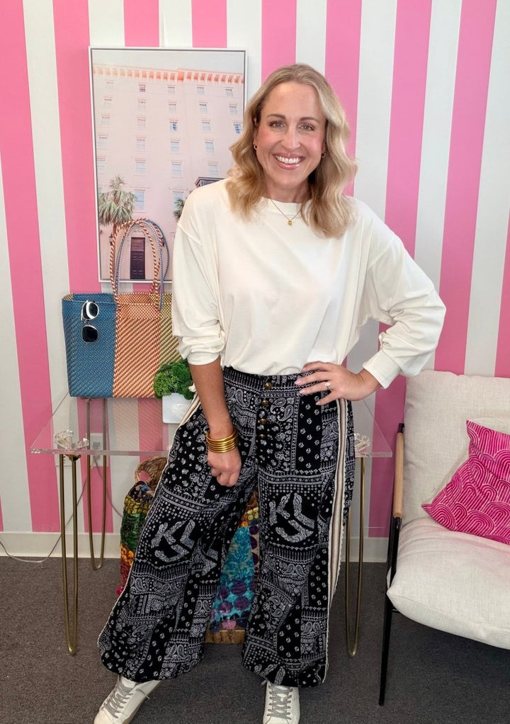 A woman posing in a white long-sleeve top and black-and-white patterned wide-leg pants, standing in front of a pink and white striped wall.