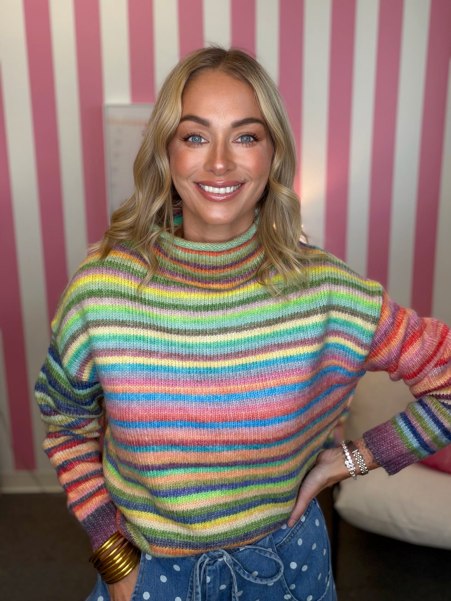 A smiling blonde woman wearing a colorful, rainbow-striped turtleneck sweater and blue polka-dot pants, standing in front of a pink and white striped wall.