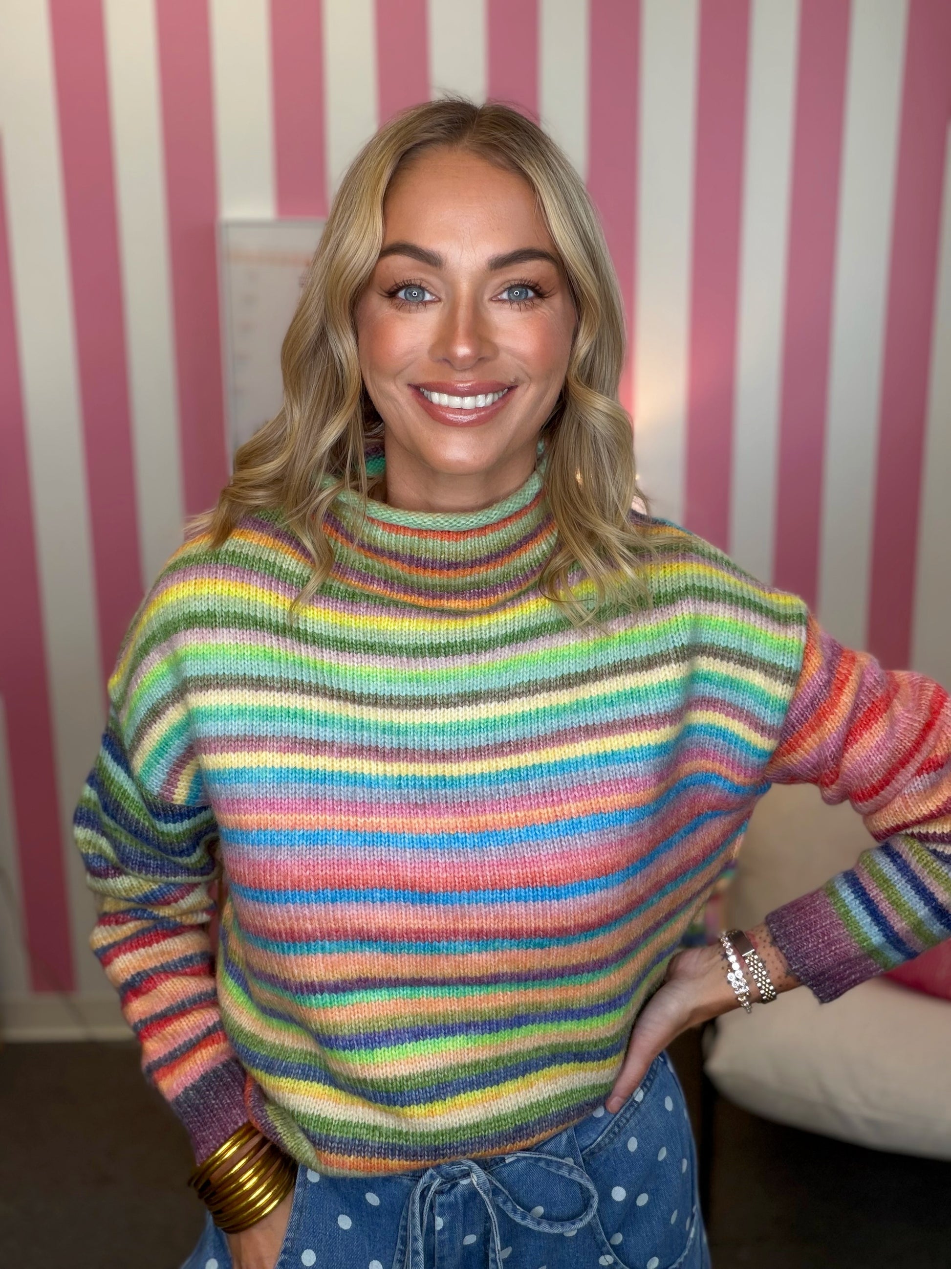A smiling blonde woman wearing a colorful, rainbow-striped turtleneck sweater and blue polka-dot pants, standing in front of a pink and white striped wall.