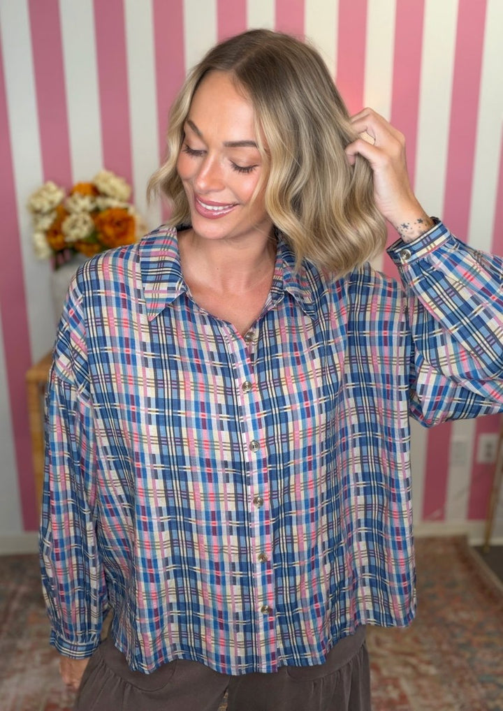 A woman wearing a blue, pink, and white plaid button-down shirt, standing in front of a pink and white striped wall.