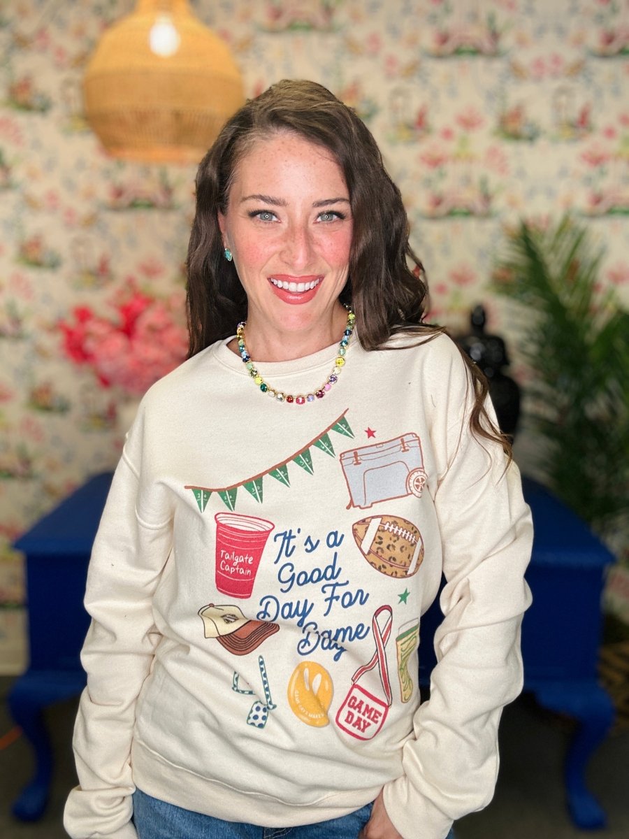 A woman smiling while wearing a cream-colored crewneck sweatshirt with a colorful graphic print that reads 'It's a Good Day For Bape' surrounded by sports and coffee-themed illustrations. The background features a floral wallpaper, a hanging lamp, and blue furniture.