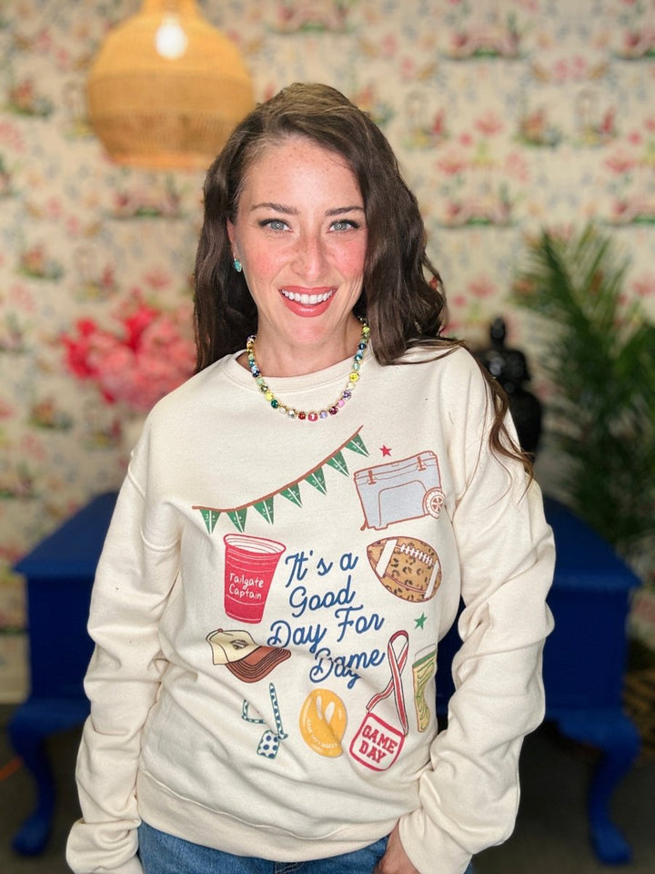 A woman smiling while wearing a cream-colored crewneck sweatshirt with a colorful graphic print that reads 'It's a Good Day For Bape' surrounded by sports and coffee-themed illustrations. The background features a floral wallpaper, a hanging lamp, and blue furniture.