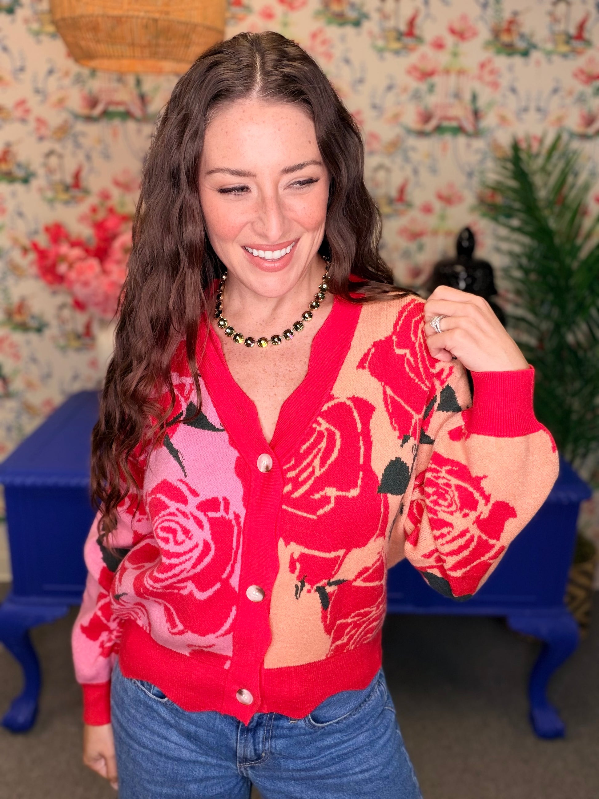 A woman wearing a red and pink cardigan with a large rose pattern, paired with blue jeans, standing in front of a floral wallpaper background.