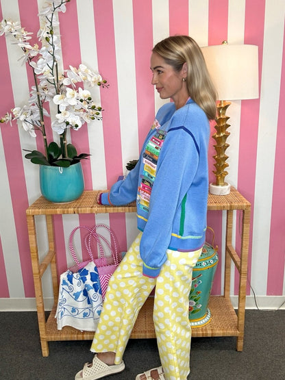 A woman stands in profile wearing a bright blue cropped zip-up jacket with colorful graphic patches and yellow-and-white polka-dot pants, posed beside a small wooden console table in a pink-and-white striped room.
