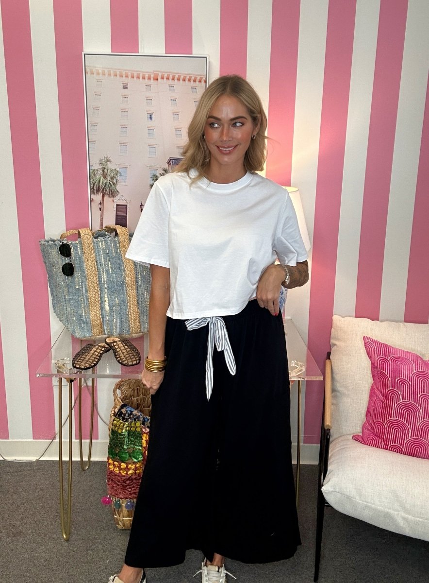 A woman stands in a room with pink and white striped walls, wearing a white short-sleeve crewneck t-shirt and a long black skirt with a white tie detail at the waist. She is standing next to a glass table displaying a blue and gold striped tote bag.