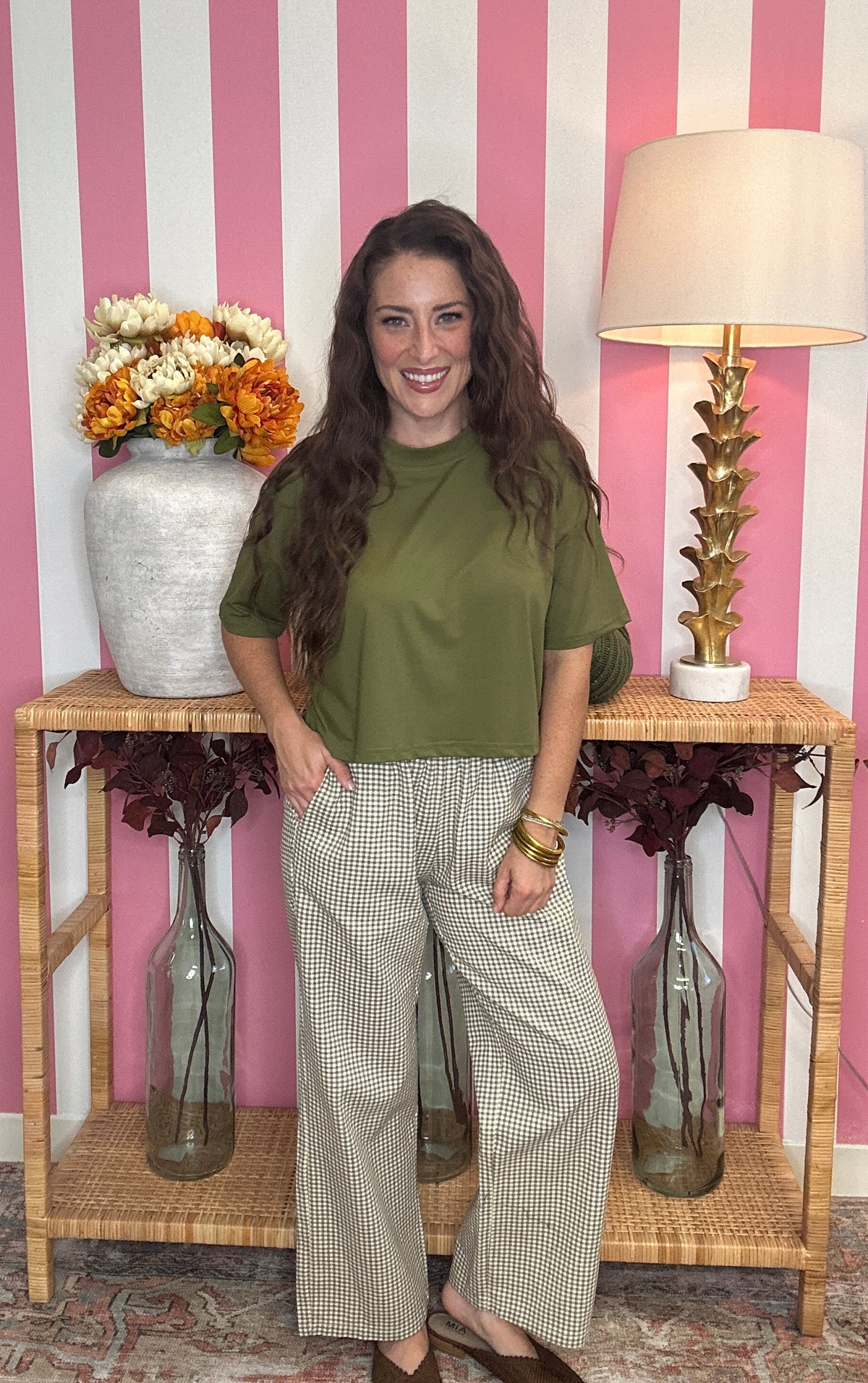 A woman stands in front of a pink and white striped wall wearing an olive green short-sleeve top and beige wide-leg houndstooth pants. She is posed in front of a wooden console table decorated with a vase of orange and white flowers, a gold lamp, and glass vases.