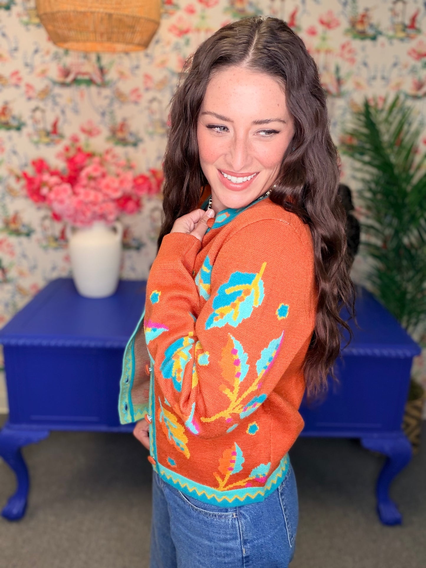 A woman wearing a bright orange knit sweater with colorful leaf and geometric patterns in blue, yellow, and green, paired with blue jeans, standing in a vintage-style room with floral wallpaper and a blue side table.
