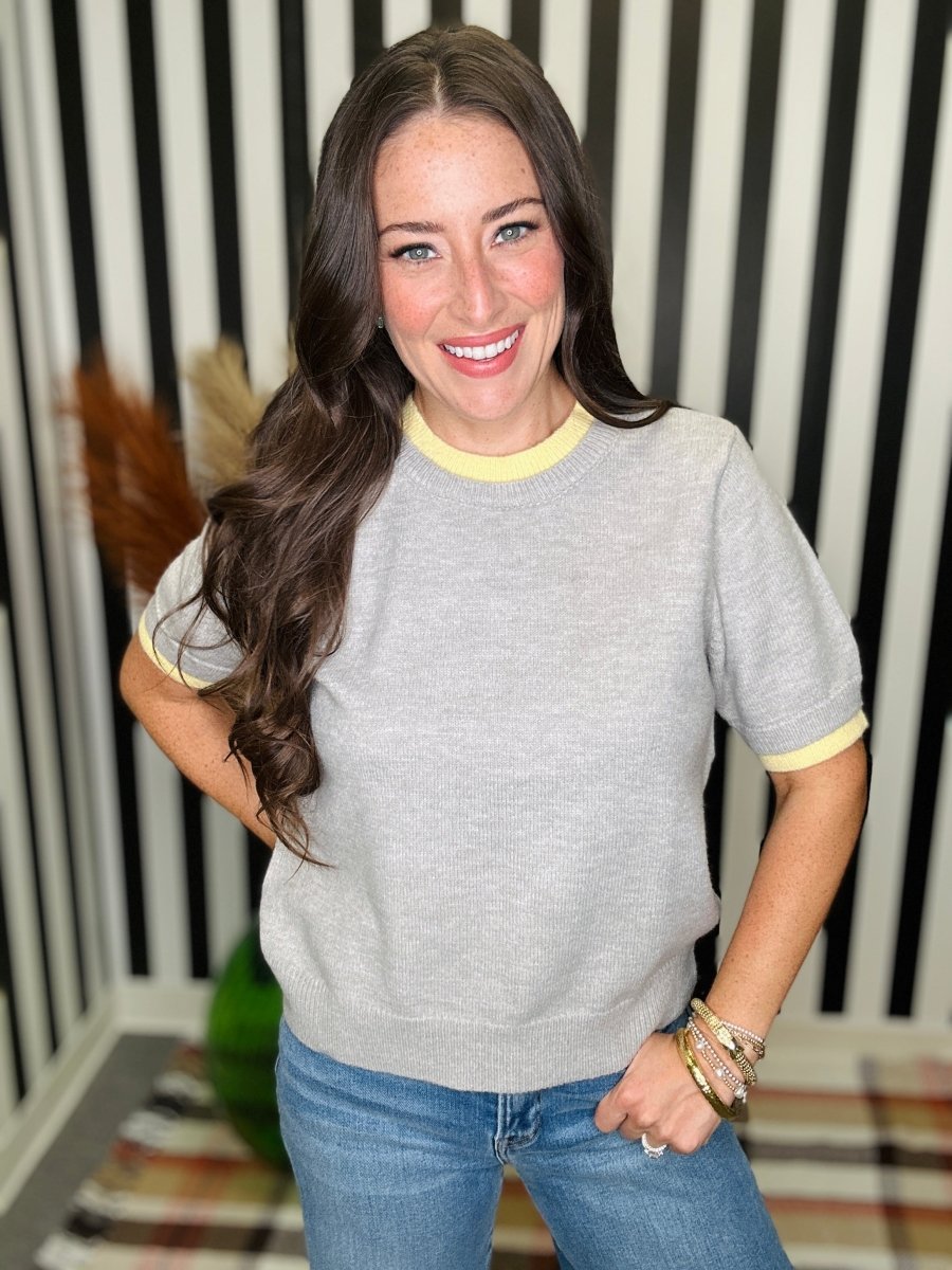 A woman smiling and wearing a light gray short-sleeve knit sweater with yellow trim on the crew neck and sleeve cuffs, paired with blue jeans, standing in front of a black and white striped wall.