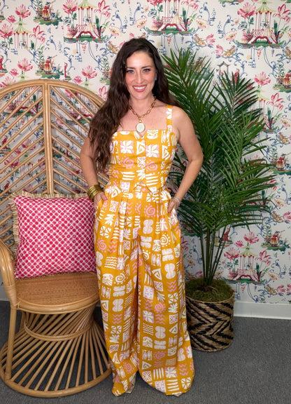 Woman in a yellow patterned jumpsuit standing in a room with floral wallpaper, a wicker chair, and a plant.