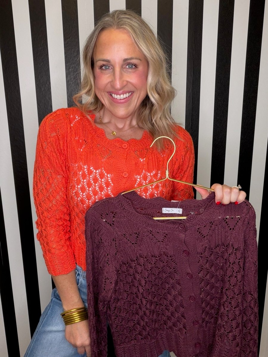 A smiling blonde woman wearing a bright orange open-knit long-sleeve top and holding a marron sweater on a hanger and blue jeans, standing against a black and white vertically striped background.