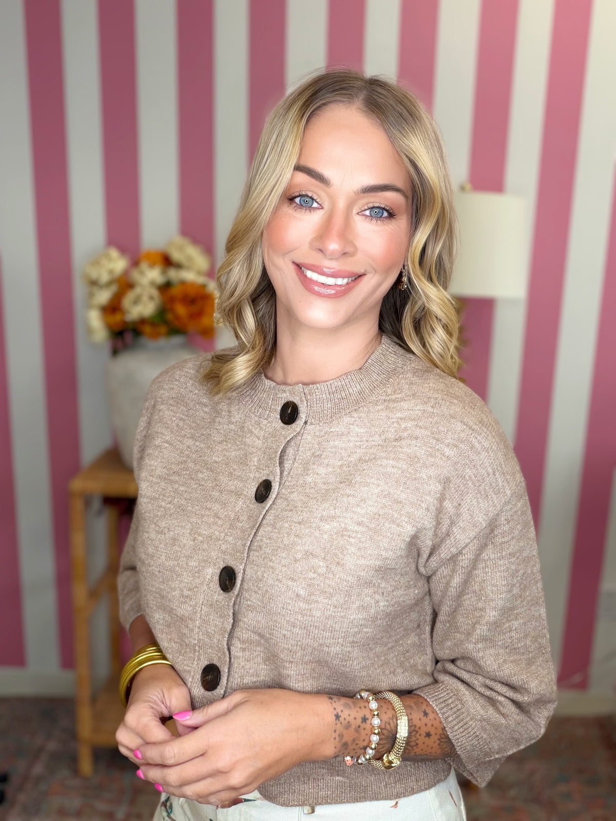 A smiling blonde woman wearing a beige knit cardigan with dark buttons, posed in front of a pink and white striped wall.