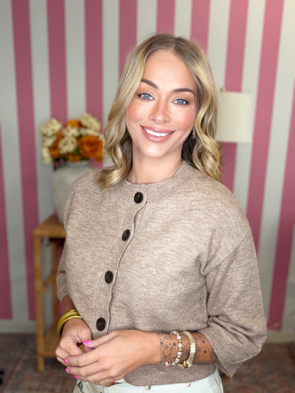 A smiling blonde woman wearing a beige knit cardigan with dark buttons, posed in front of a pink and white striped wall.