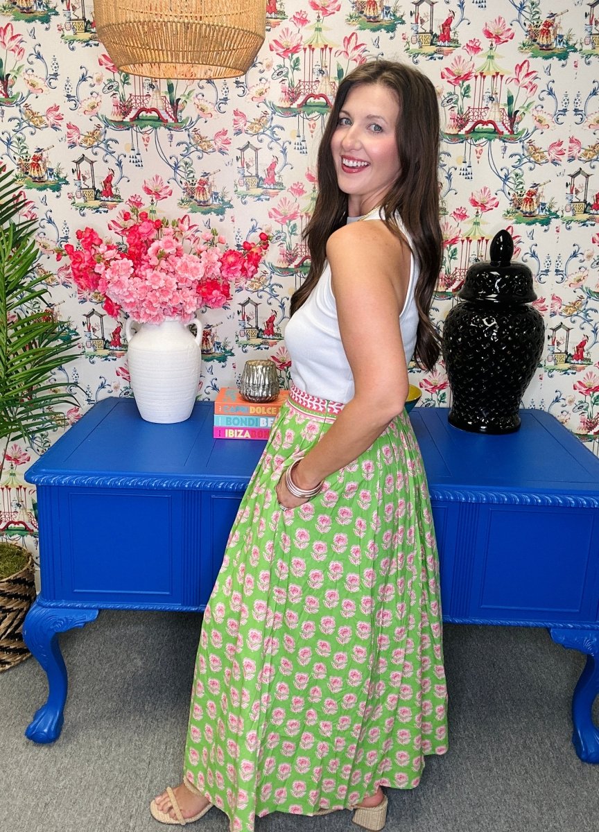 Woman in a green and pink patterned dress standing in a room with floral wallpaper and blue tables.
