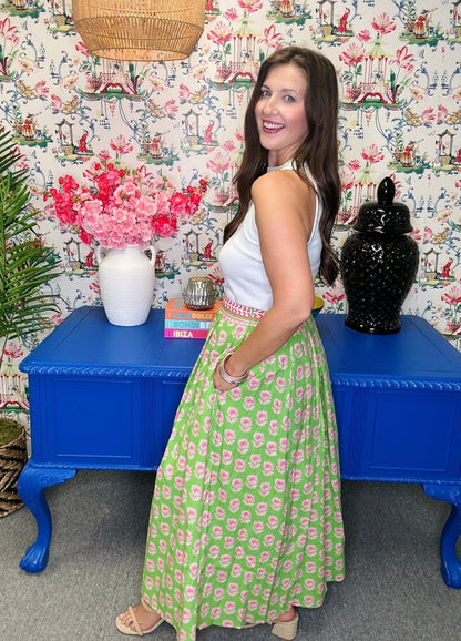 Woman in a green and pink patterned dress standing in a room with floral wallpaper and blue tables.