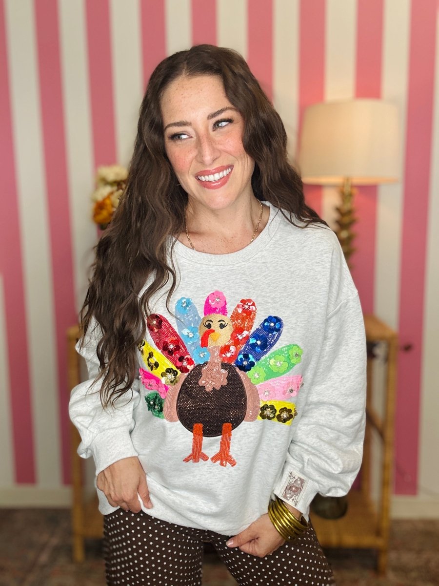 A woman smiling while wearing a light gray crewneck sweatshirt with a colorful sequined turkey graphic on the front, paired with brown polka dot pants, set against a pink and white striped wall background.