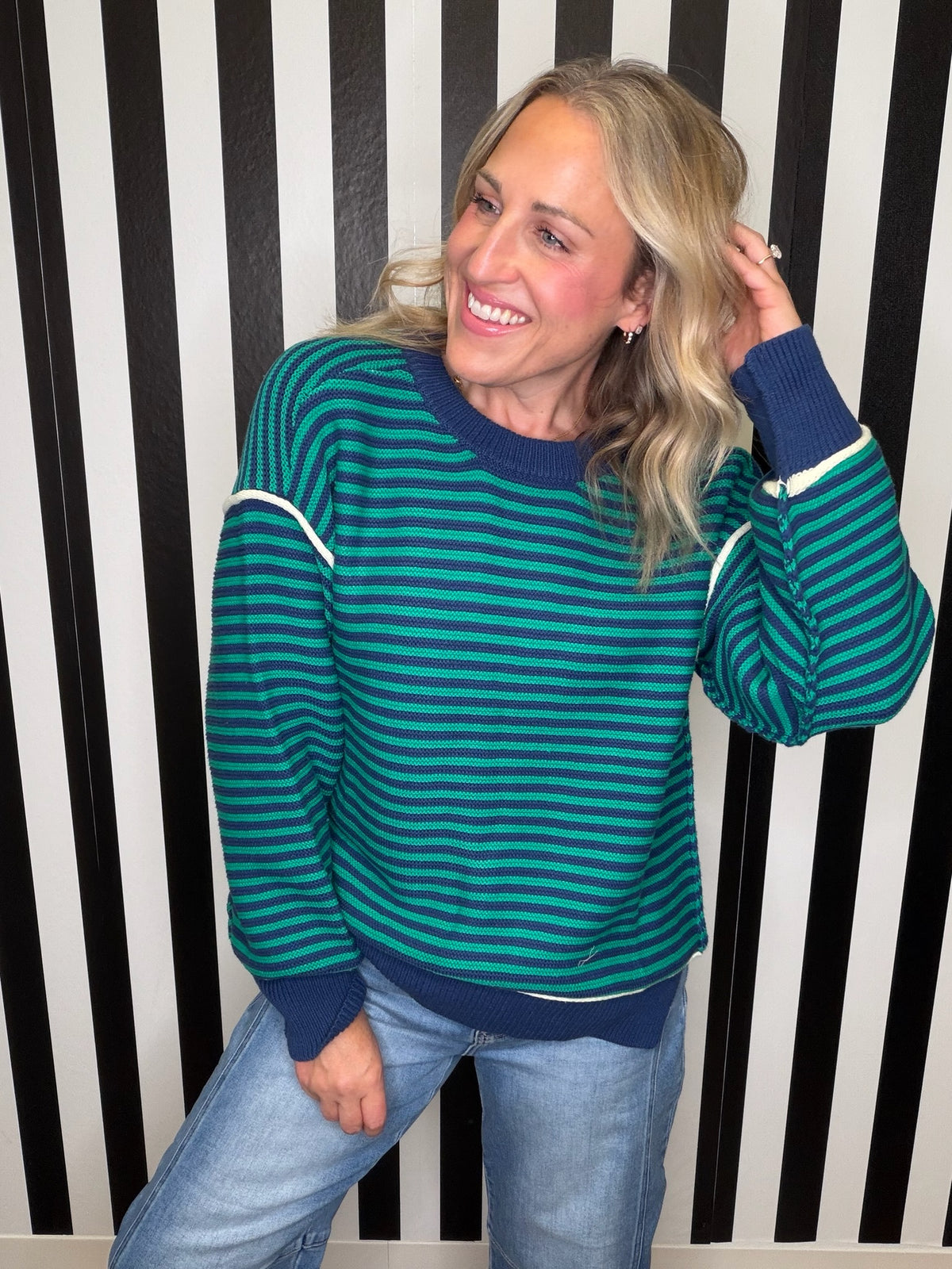 A woman wearing a teal and green striped long-sleeve sweater with navy blue ribbed trim, paired with light blue jeans, standing in front of a black and white vertically striped wall.