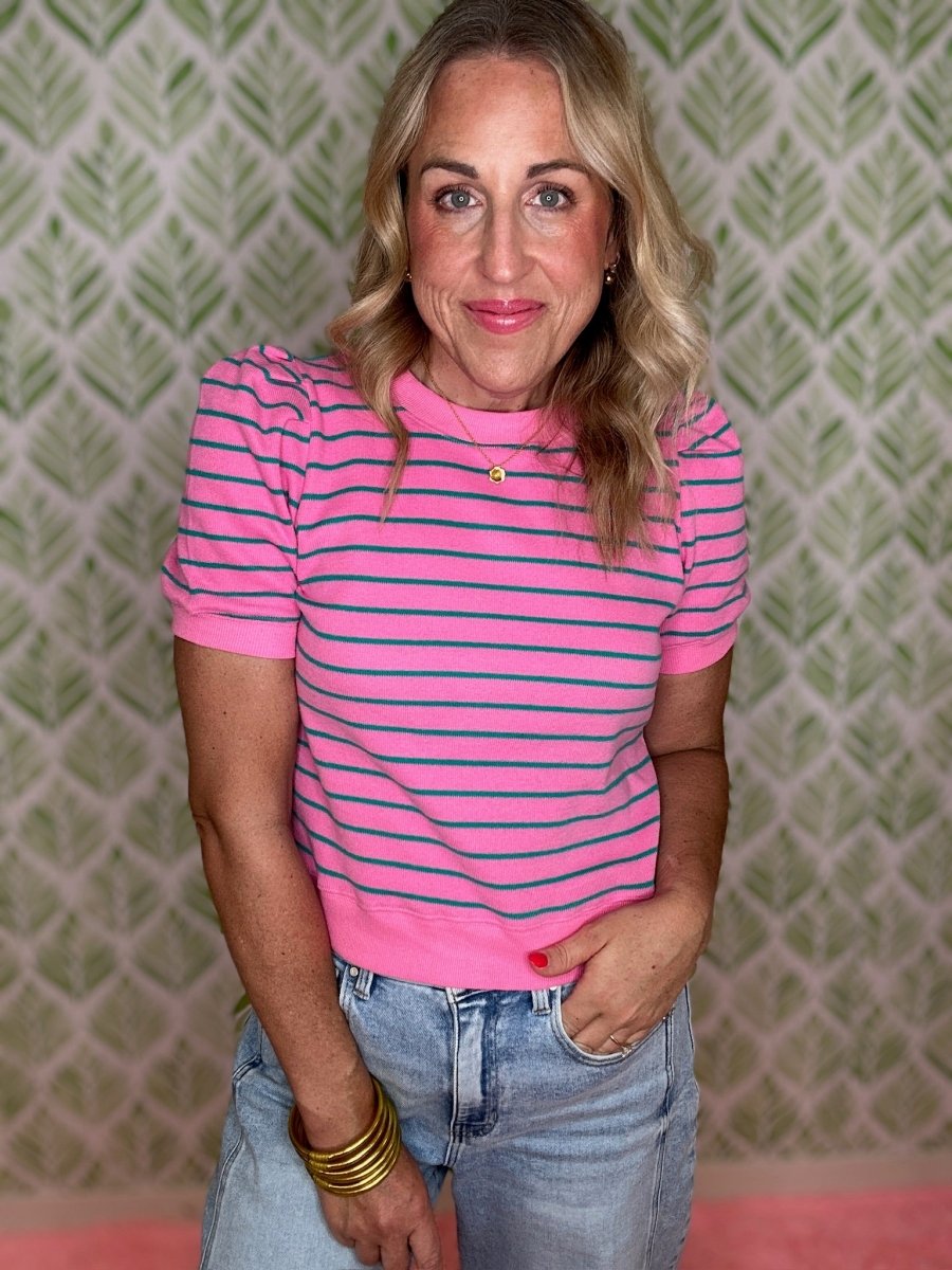 A woman wearing a short-sleeve pink knit top with thin dark blue horizontal stripes, paired with light-wash blue jeans and gold bracelets, standing in front of a green and white patterned wallpaper background.
