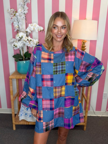 A woman wearing a blue patchwork plaid tunic dress, standing in front of a pink and white striped wall with a lamp and a side table.