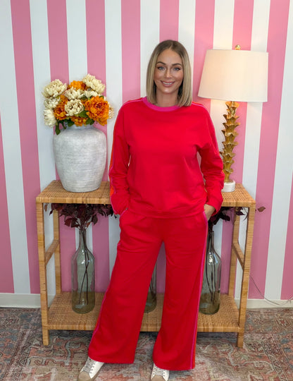 A woman modeling a bright red two-piece lounge set consisting of a long-sleeve sweatshirt and matching wide-leg pants, standing in front of a pink and white striped wall.