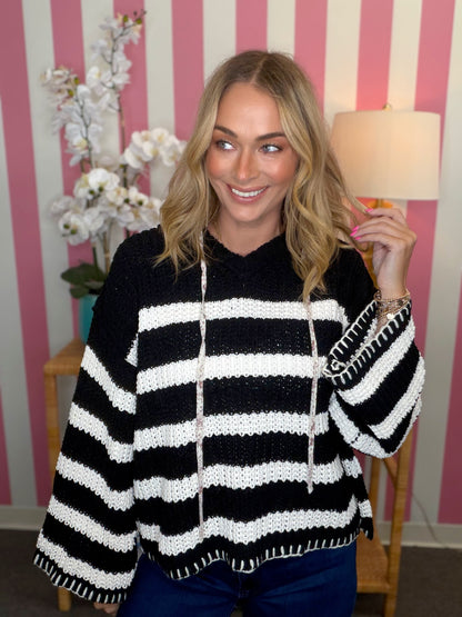 A woman wearing a black and white striped knit hoodie sweater, standing in front of a pink and white striped wall with a lamp and flowers in the background.