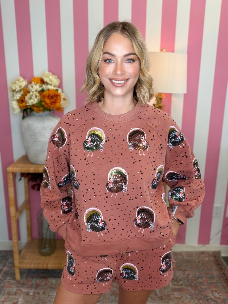 A woman modeling a matching dusty rose sweatshirt and shorts set with a black and white skull print, set against a pink and white striped wall background.