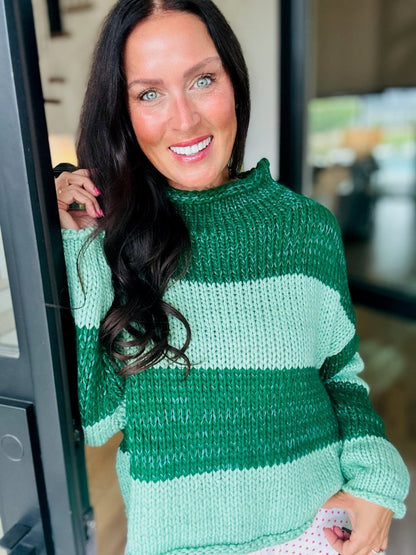 A smiling woman with black hair wearing a chunky knit sweater with green and mint green geometric blocks.