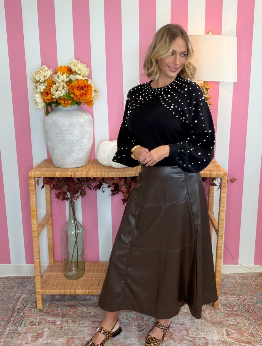 A woman stands indoors wearing a black long-sleeve top with silver rhinestone embellishments and a dark brown faux-leather midi skirt. She is posed in front of a pink and white striped wall with a wicker console table holding a vase of orange and white flowers.