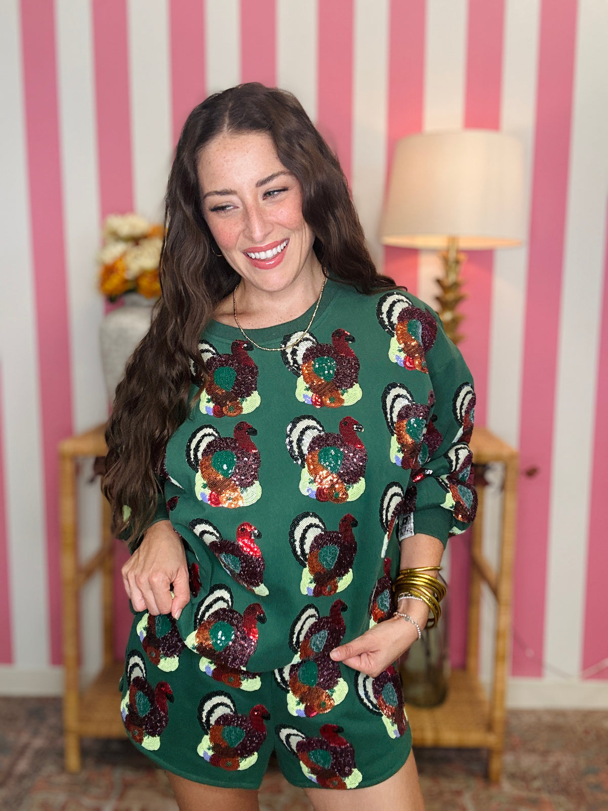 A woman wearing a green sequined romper with a repeating rooster pattern, standing in front of a pink and white striped wall with a lamp and wooden furniture in the background.