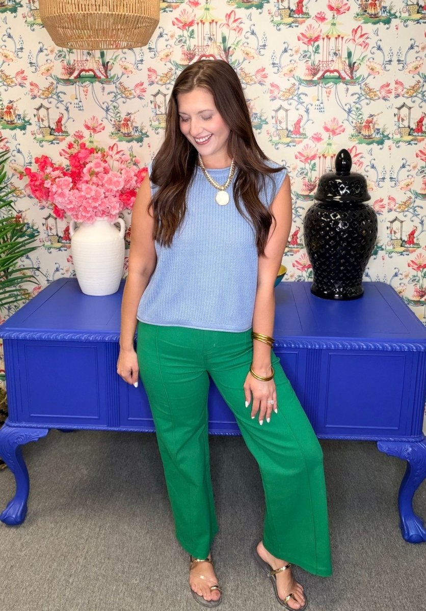 Woman in a blue top and green pants standing in front of a colorful floral-patterned wall.