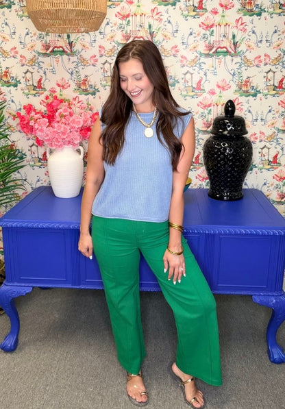 Woman in a blue top and green pants standing in front of a colorful floral-patterned wall.