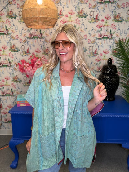 A woman wearing a light mint green patterned shawl over a white top and blue jeans, standing in front of a blue console table and floral wallpaper.