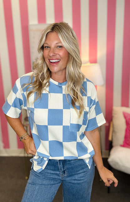 A woman with blonde hair smiling and wearing a blue and white checkered short-sleeve top with blue jeans, standing in front of a pink and white striped wall.