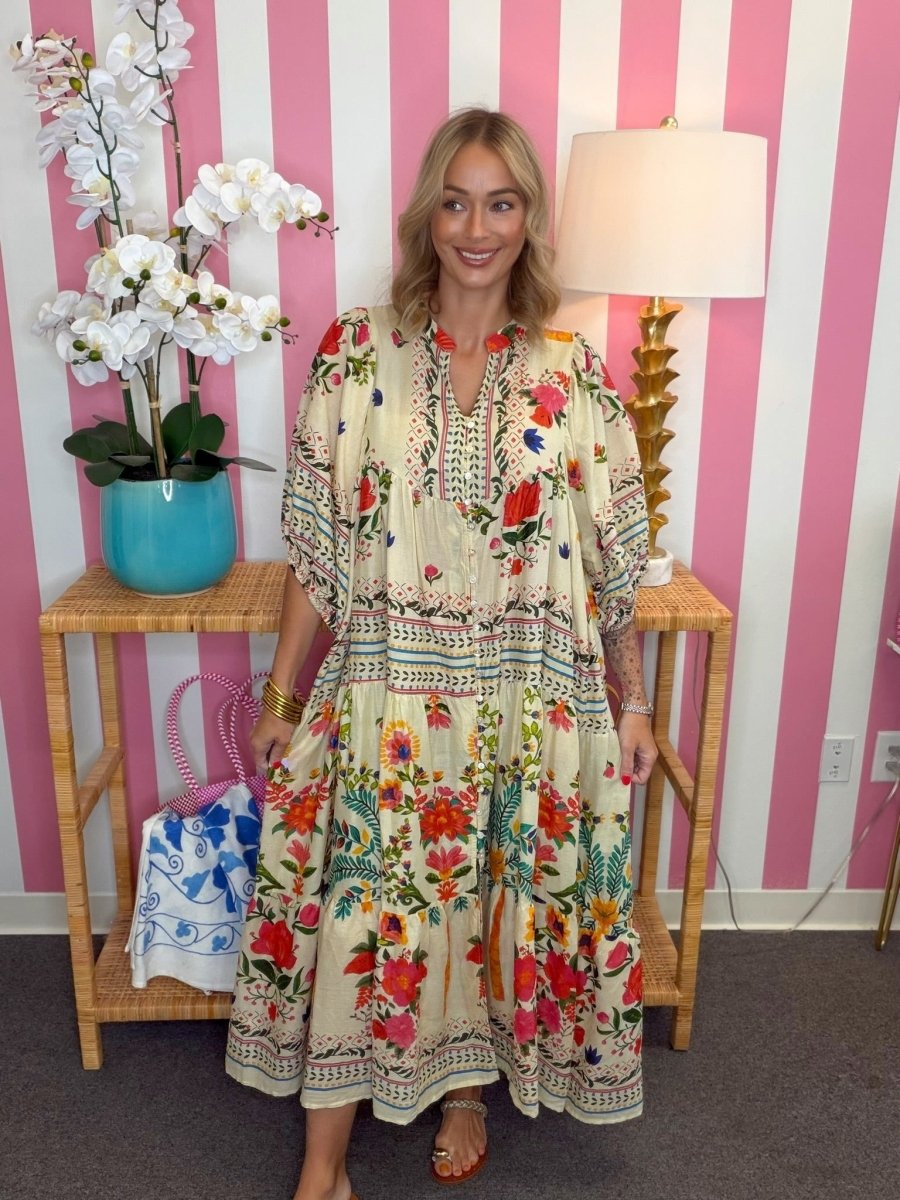 A woman modeling a bohemian-style tiered maxi dress with a cream base and a vibrant red, green, and blue floral print, standing in front of a pink and white striped wall.