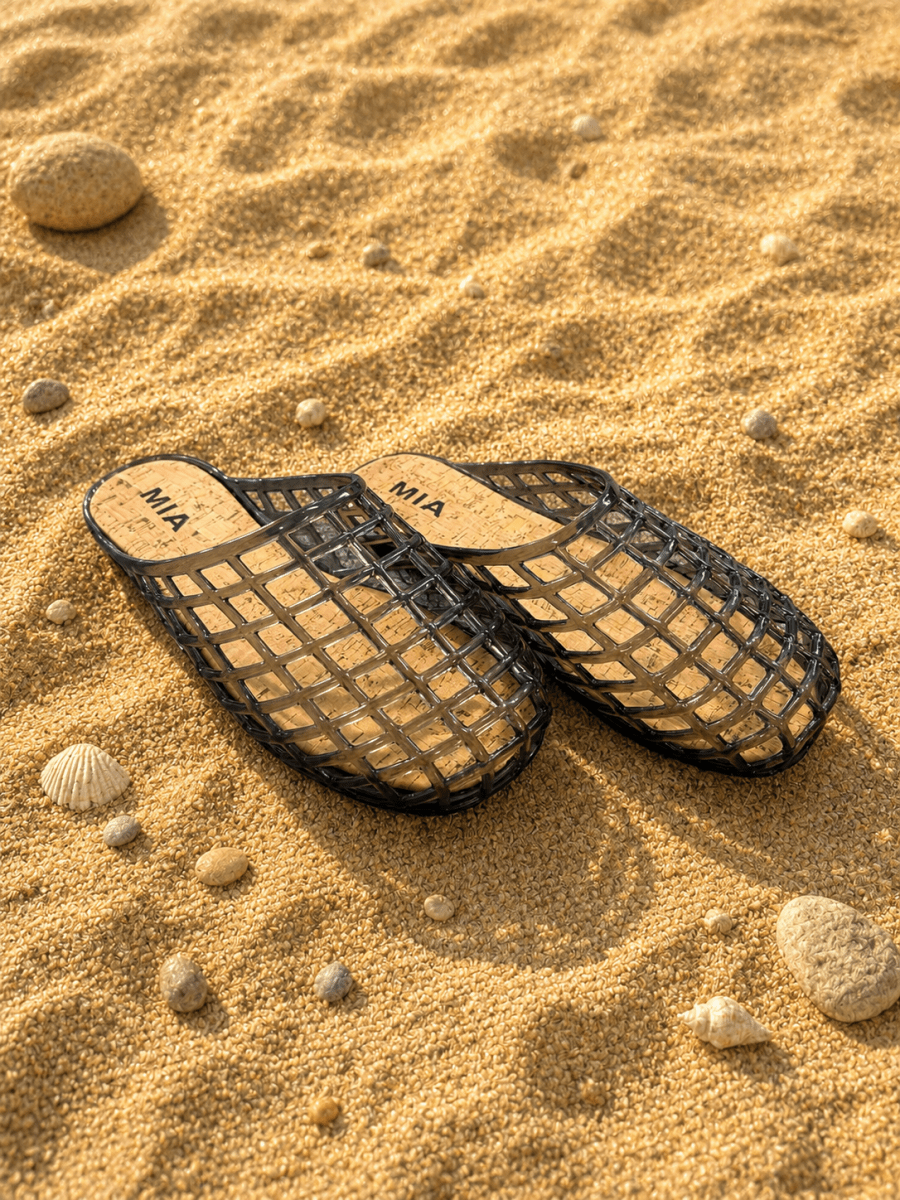 Black sandals with 'MIA' branding on a sandy surface with shells and rocks.