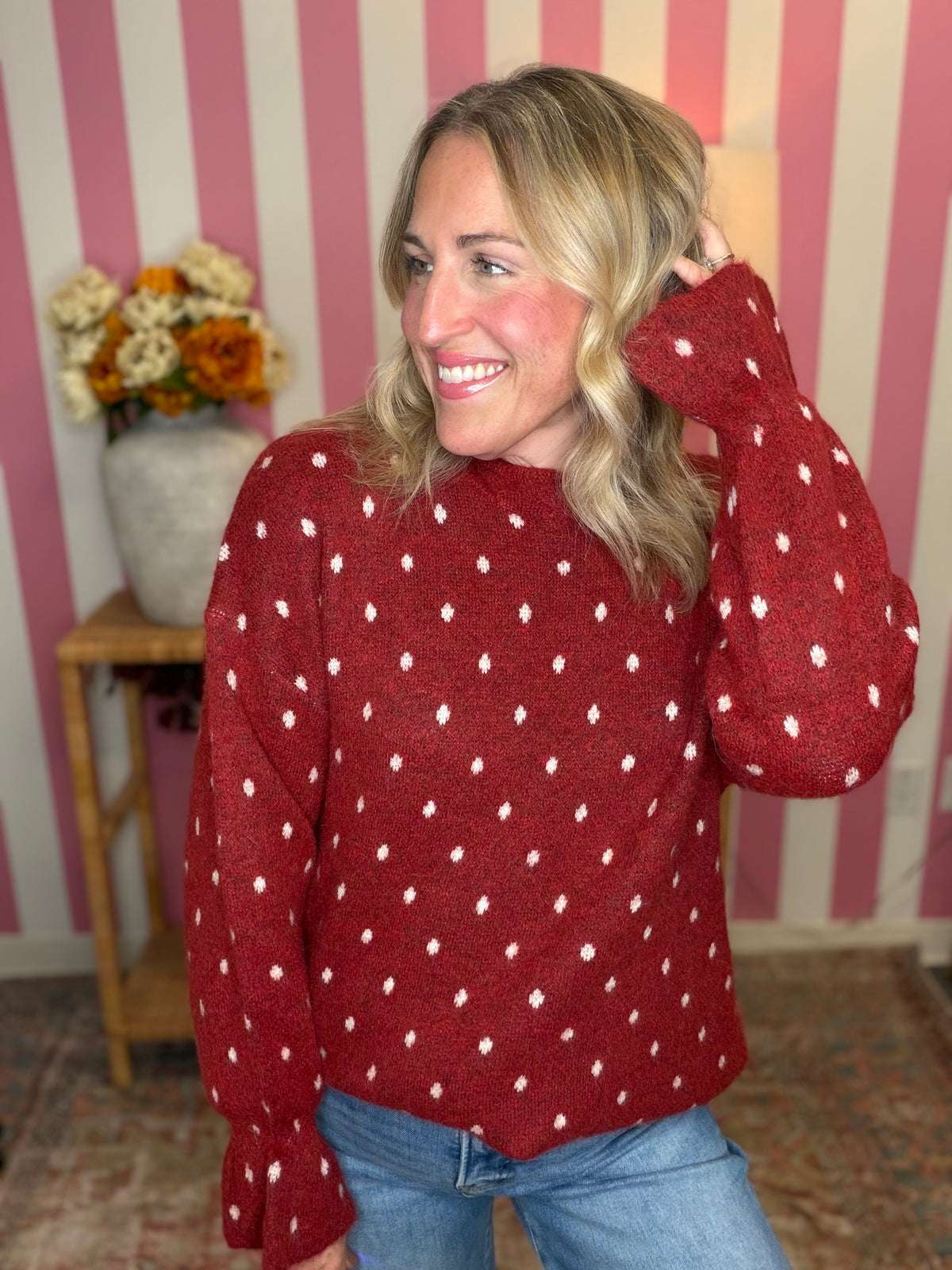 A woman wearing a red knit sweater with a white geometric dot pattern, paired with blue jeans, standing in front of pink and white striped wallpaper.