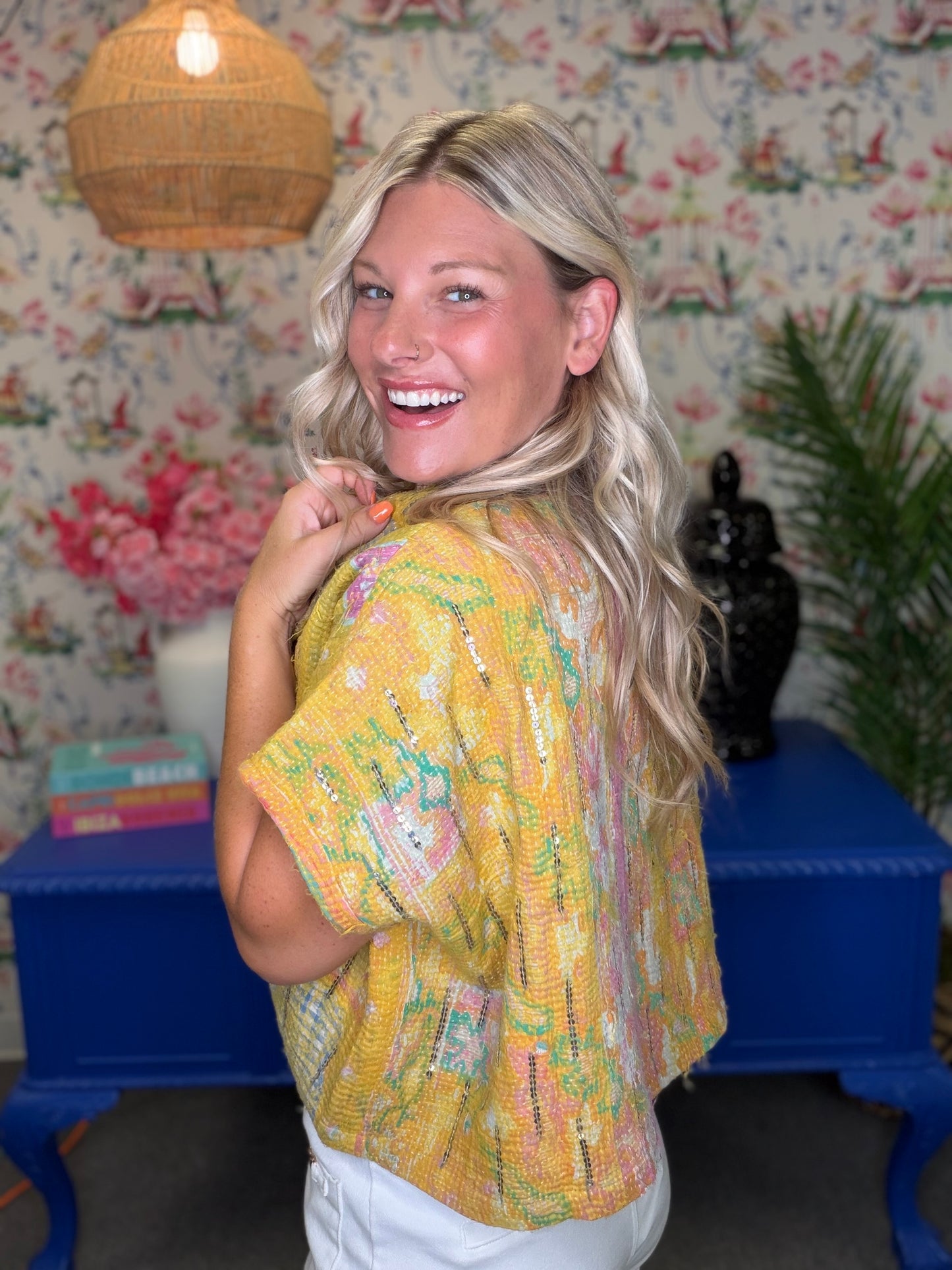 A smiling blonde woman wearing a yellow short-sleeve knit top with multicolored abstract patterns, posed in a decorated room with floral wallpaper, a blue table, and a wicker pendant light.