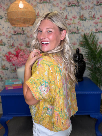 A smiling blonde woman wearing a yellow short-sleeve knit top with multicolored abstract patterns, posed in a decorated room with floral wallpaper, a blue table, and a wicker pendant light.