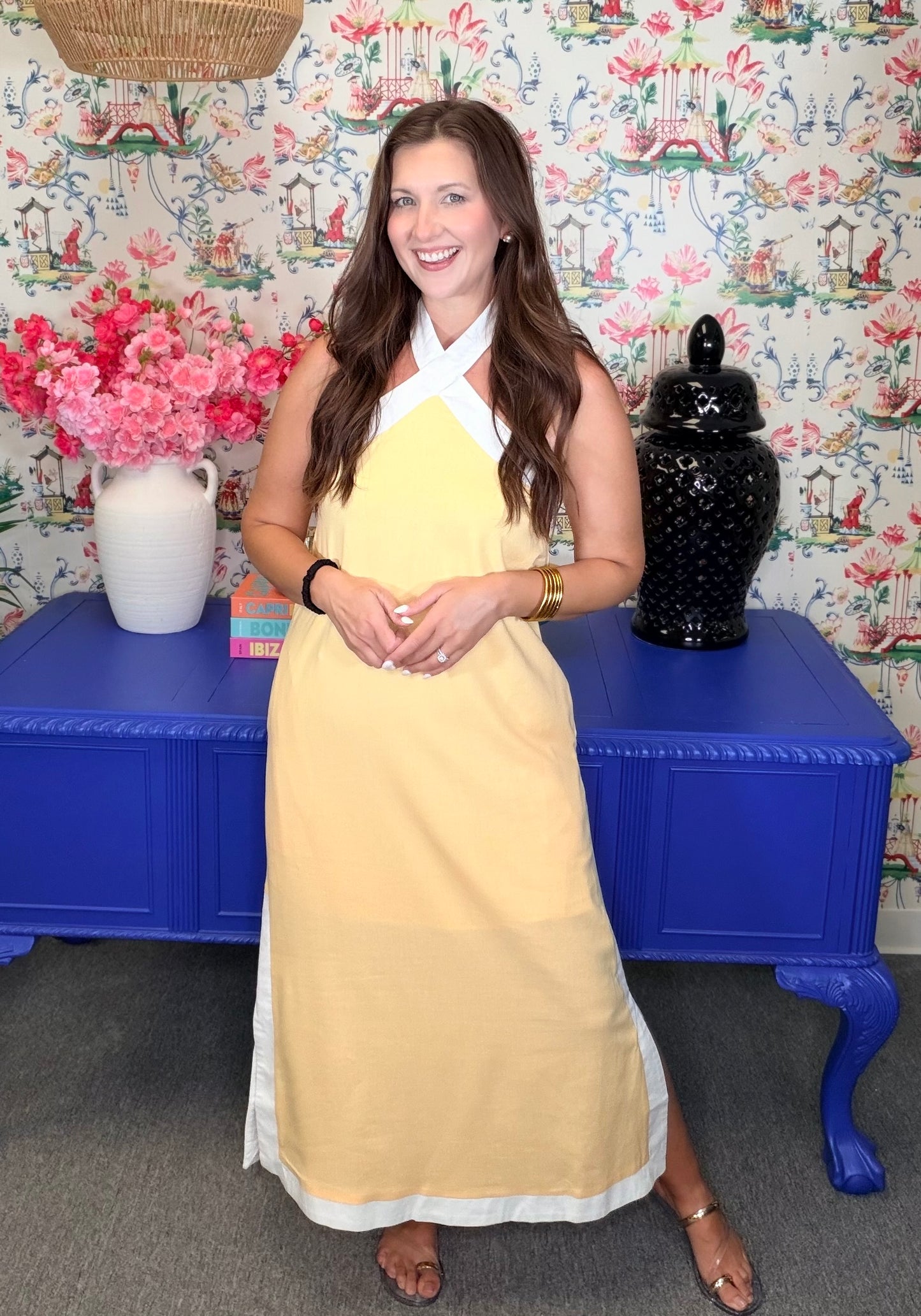 Woman in a yellow dress standing in front of a colorful wall with floral patterns and decorative items.