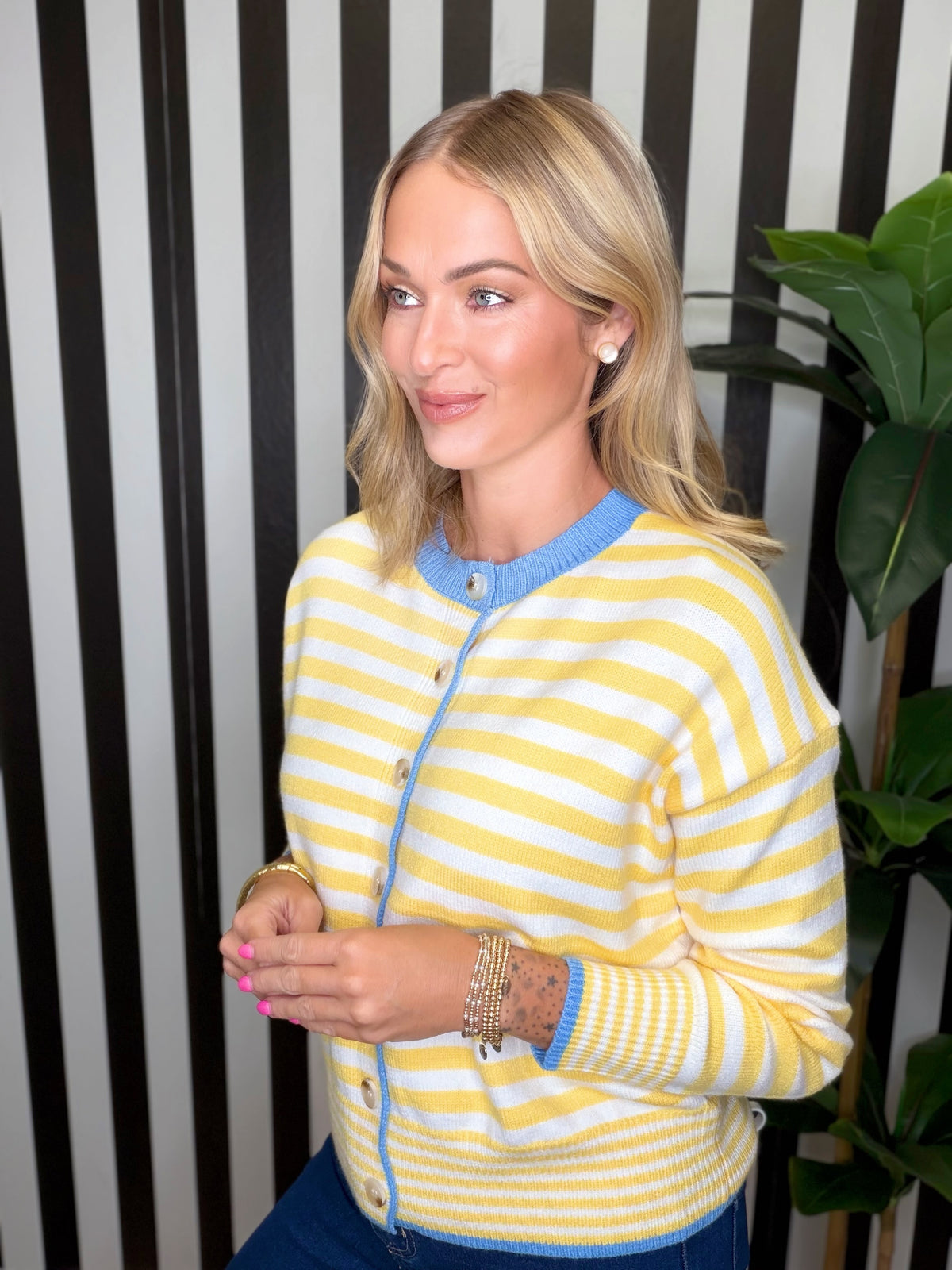 A woman wearing a yellow and white striped knit cardigan with light blue trim, standing in front of a black and white striped wall with a green plant on the right.