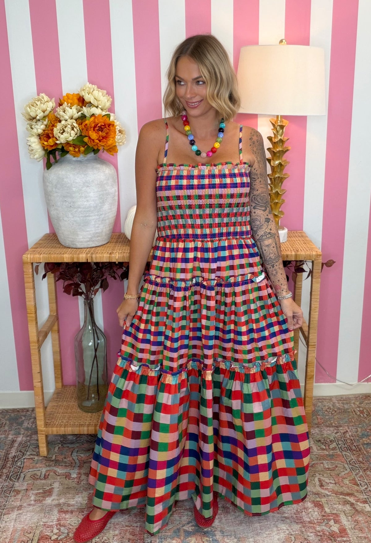 A woman models a colorful, tiered maxi dress with a vibrant multicolor plaid pattern. She is standing in front of a pink and white striped wall, with a wooden side table holding a vase of flowers and a lamp in the background.
