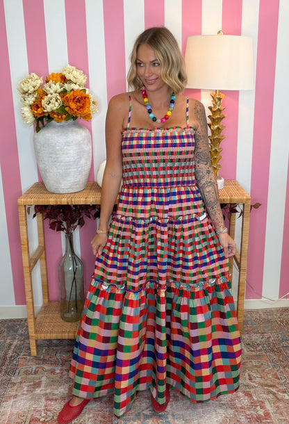 A woman models a colorful, tiered maxi dress with a vibrant multicolor plaid pattern. She is standing in front of a pink and white striped wall, with a wooden side table holding a vase of flowers and a lamp in the background.