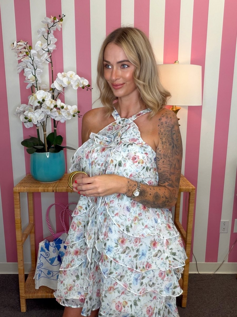 A woman wearing a white halter-neck ruffled dress with a pink, blue, and green floral print, standing in front of a pink and white striped wall next to a wooden side table with a turquoise orchid pot.
