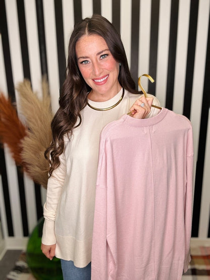A woman with long brown hair smiles while holding up a light pink long-sleeve knit top on a gold hanger. She is wearing an off-white long-sleeve top and blue jeans, standing in front of a black and white striped wall.