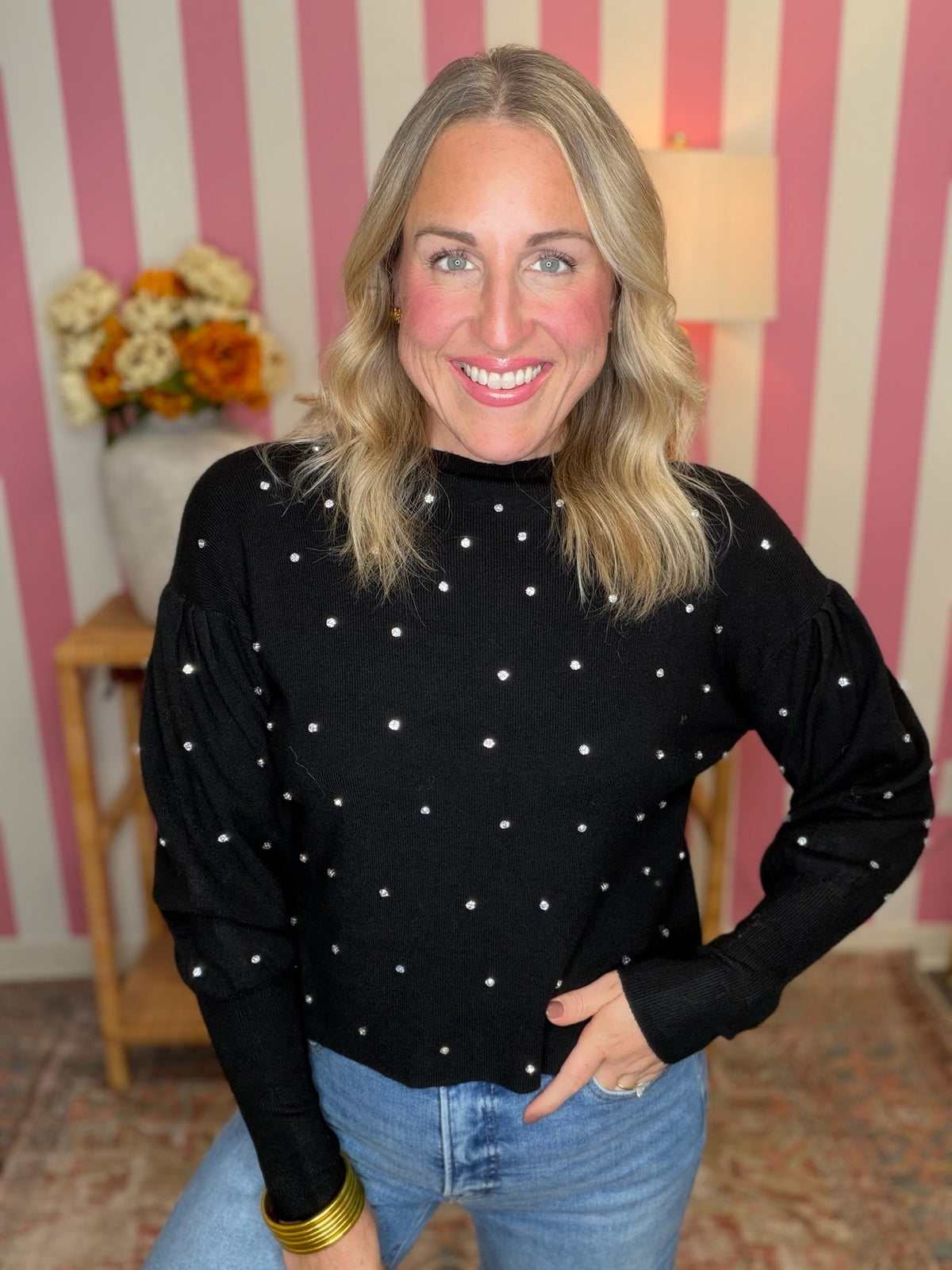 A smiling blonde woman wearing a black long-sleeve mock neck top with small white polka dots, paired with blue jeans, standing in front of pink and white striped wallpaper.