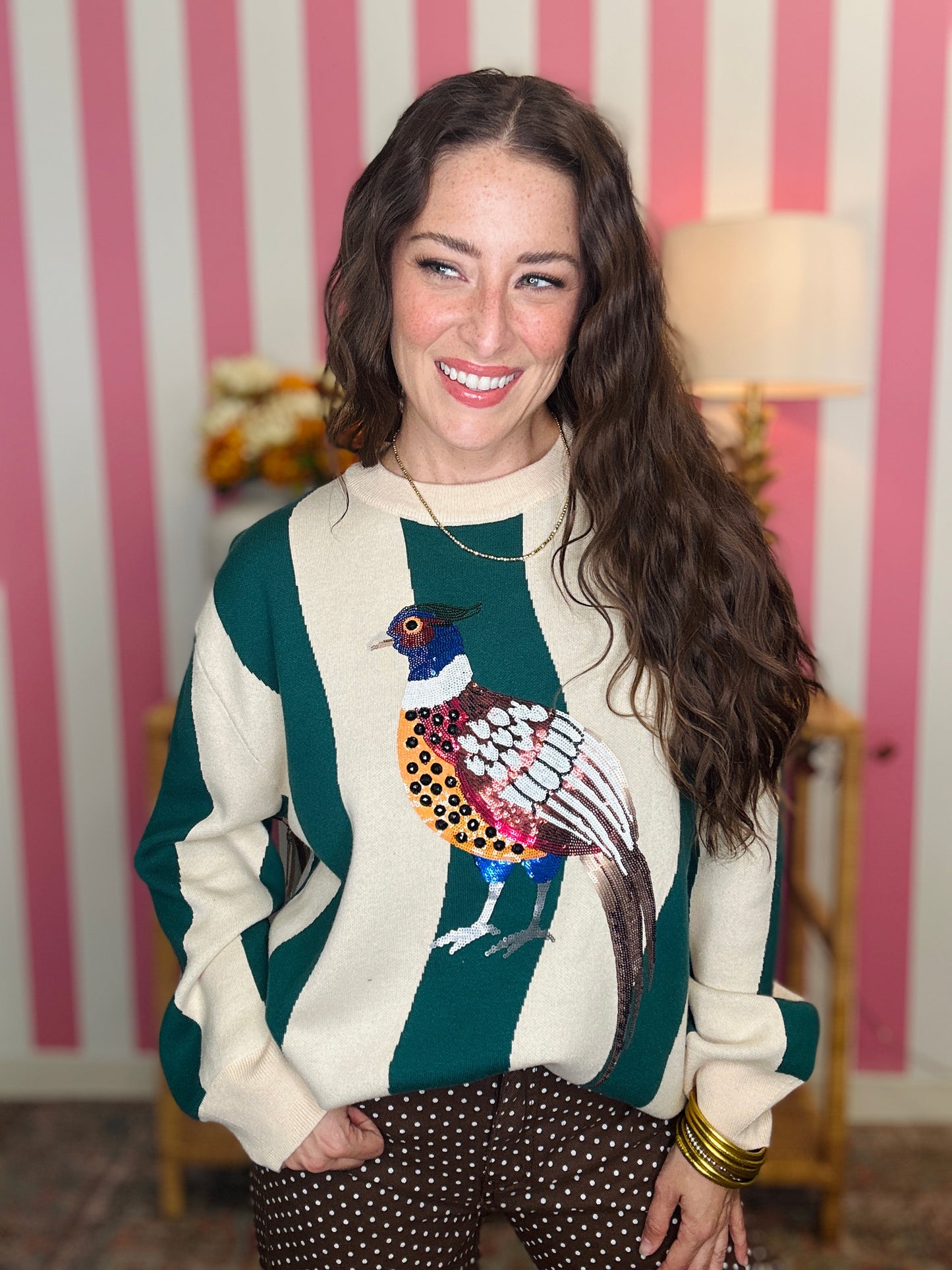 A woman smiling while wearing a cream and dark green striped sweater with a colorful peacock graphic on the front, paired with brown polka dot pants, set against a pink and white striped wall background.